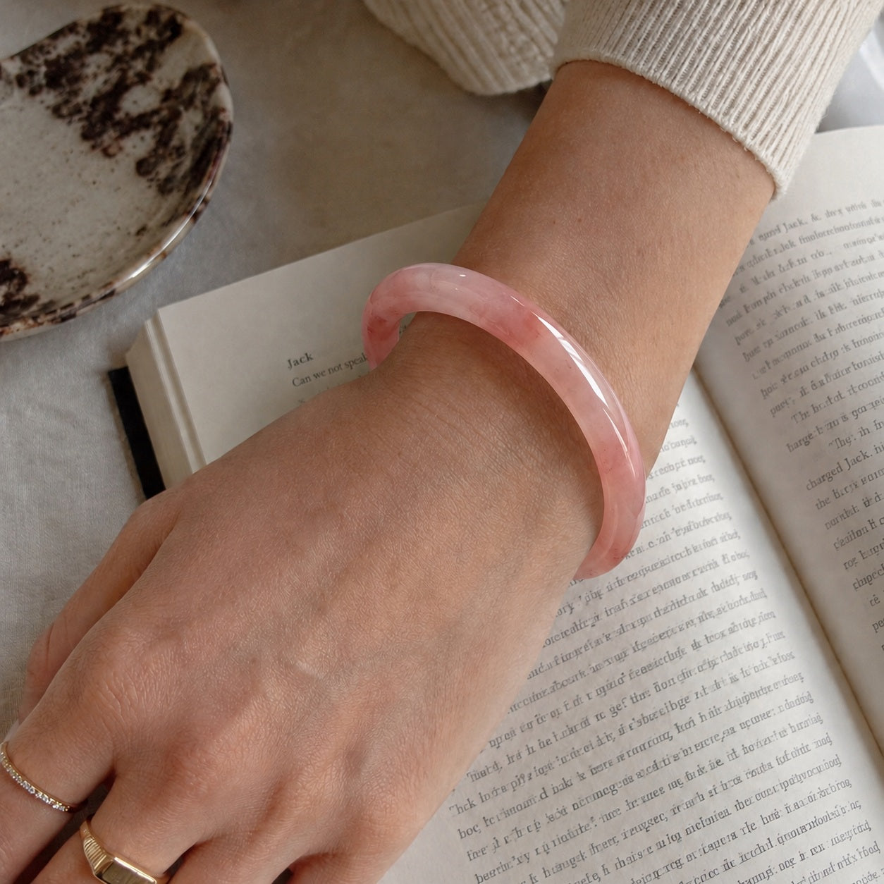 Close-up of a pink quartzite jade bangle bracelet worn on the wrist beside an open book