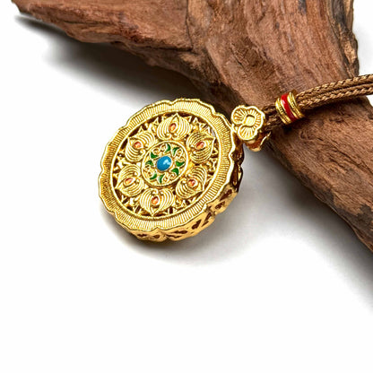 A Tibetan-style sand gold amulet pendant featuring a prominent green center stone, resting on natural driftwood background.