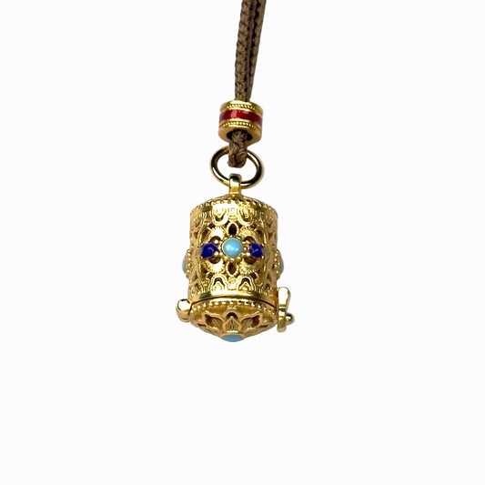 A front closed view of an authentic sand gold cylindrical prayer box pendant featuring intricate filigree openwork and colorful enamel gemstone accents on a white background.